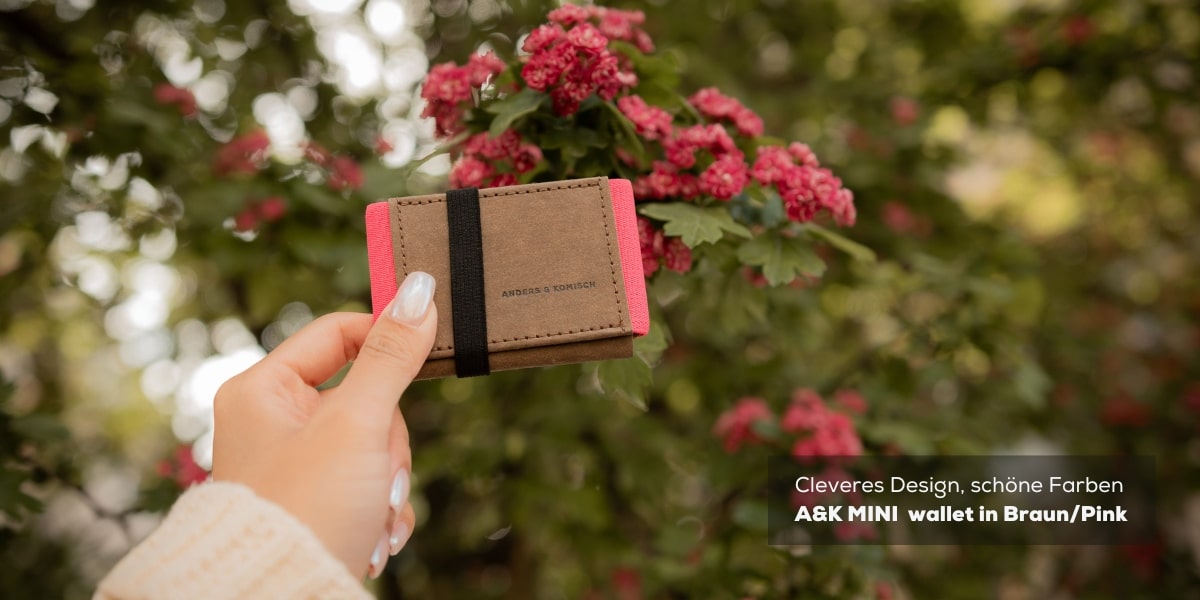 Ein A&K MINI Portemonnaie (slim wallet) in Braun/Pink, gehalten von einer Frau mit schönen langen Fingernägeln. Im Hintergrund sind zarte rosa Blumen zu sehen, die die feminine Eleganz des Produkts unterstreichen.